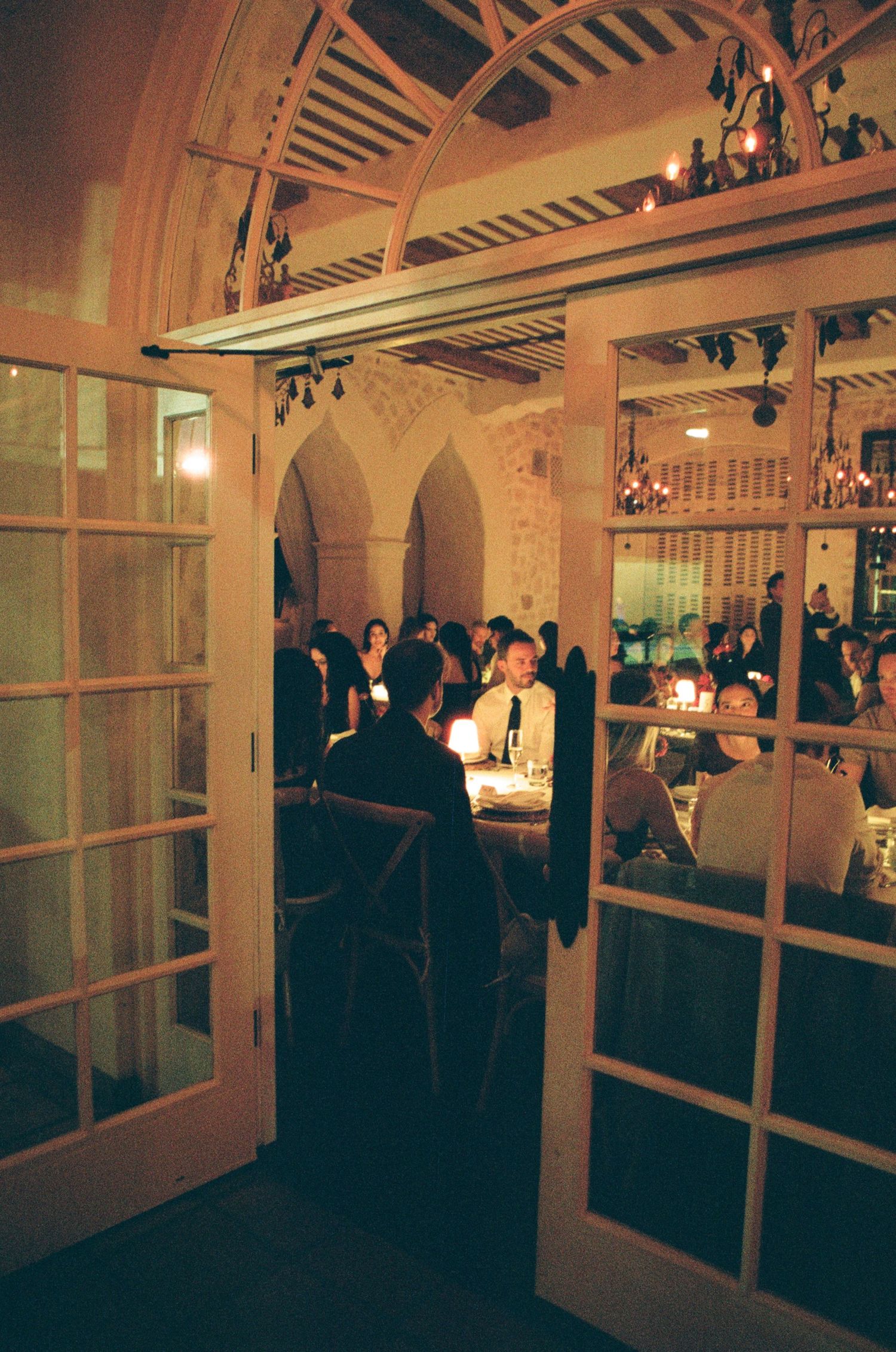 Elegant candlelit dinner party inside a rustic restaurant with arched ceilings, French doors, and guests dining by warm ambient light.