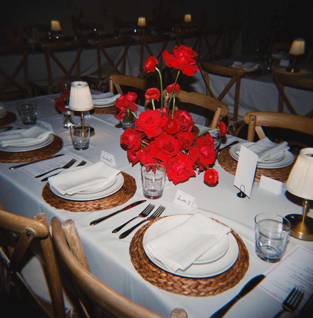 Elegant wedding table setting with red roses centerpiece, wicker chargers, white plates, and black cutlery on white tablecloth.