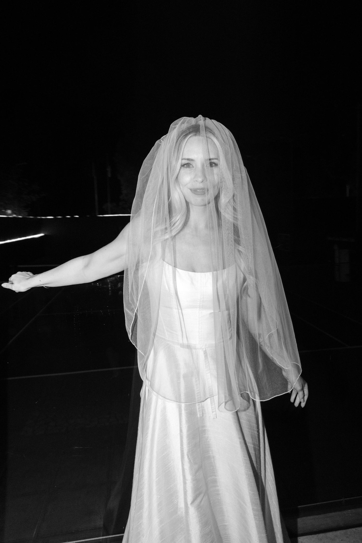 Black and white photo of a bride in a satin gown and veil, posing elegantly with one arm extended.