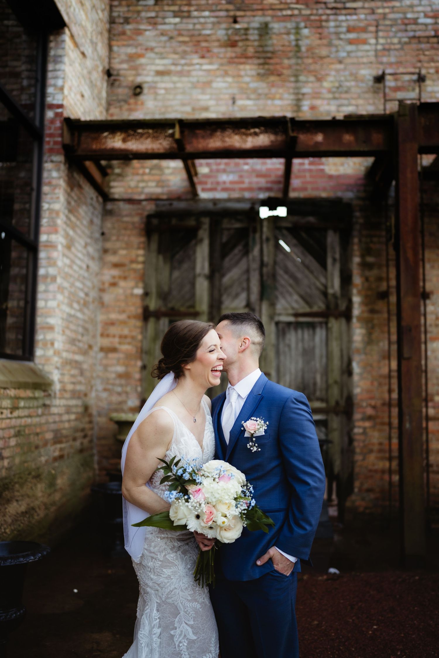 Bride and groom share a romantic moment in front of a rustic brick wall, bride holding a pastel bouquet.