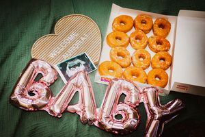 Baby shower celebration with pink BABY balloons, a box of glazed donuts, ultrasound photo, and a heart-shaped welcome sign.