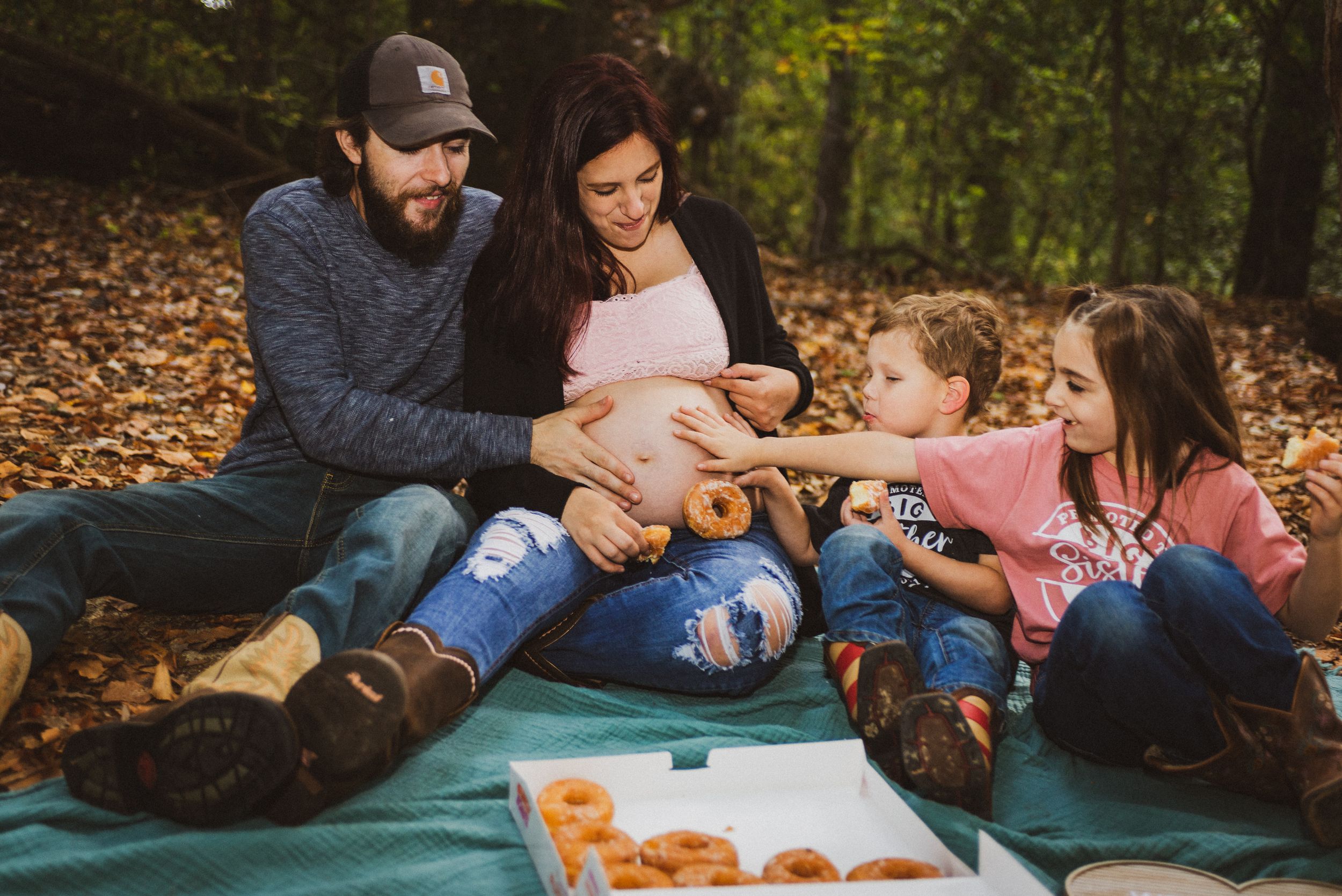 Pregnant woman enjoys donuts with family on a picnic blanket in autumn woods with two children and partner.