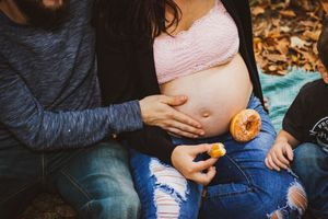 Pregnant woman sitting outdoors with partner's hand on baby bump, holding a donut, surrounded by autumn leaves.