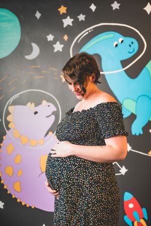Pregnant woman in floral dress cradling baby bump, smiling in front of colorful space dinosaur mural.