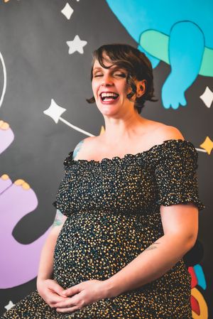 Laughing pregnant woman in black floral off-shoulder dress poses in front of colorful mural with stars.