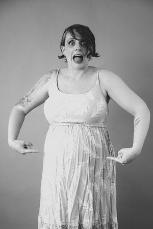 Surprised pregnant woman in floral dress pointing at baby bump, black and white studio portrait photo.