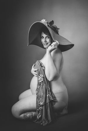 Black and white maternity photo of pregnant woman wearing wide-brim hat, holding floral fabric while posing gracefully.