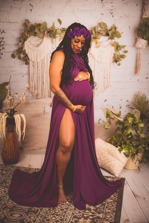 Pregnant woman in purple maternity gown with floral crown poses in boho studio with macrame and greenery.