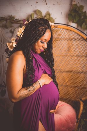 Pregnant woman in purple dress cradling baby bump, seated in wicker chair with floral backdrop.