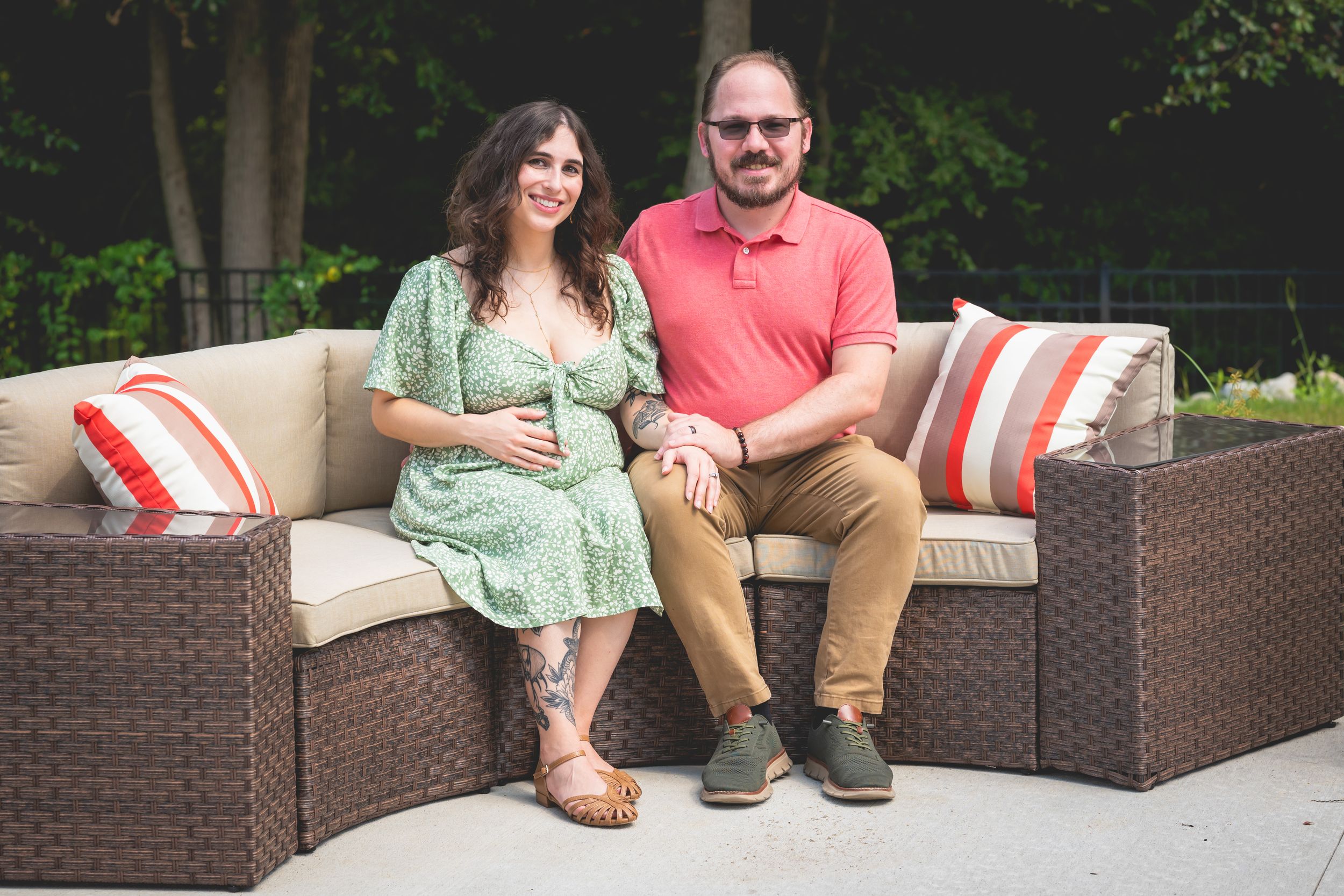 Pregnant woman in green floral dress sits with man in pink shirt on outdoor patio furniture, smiling together.