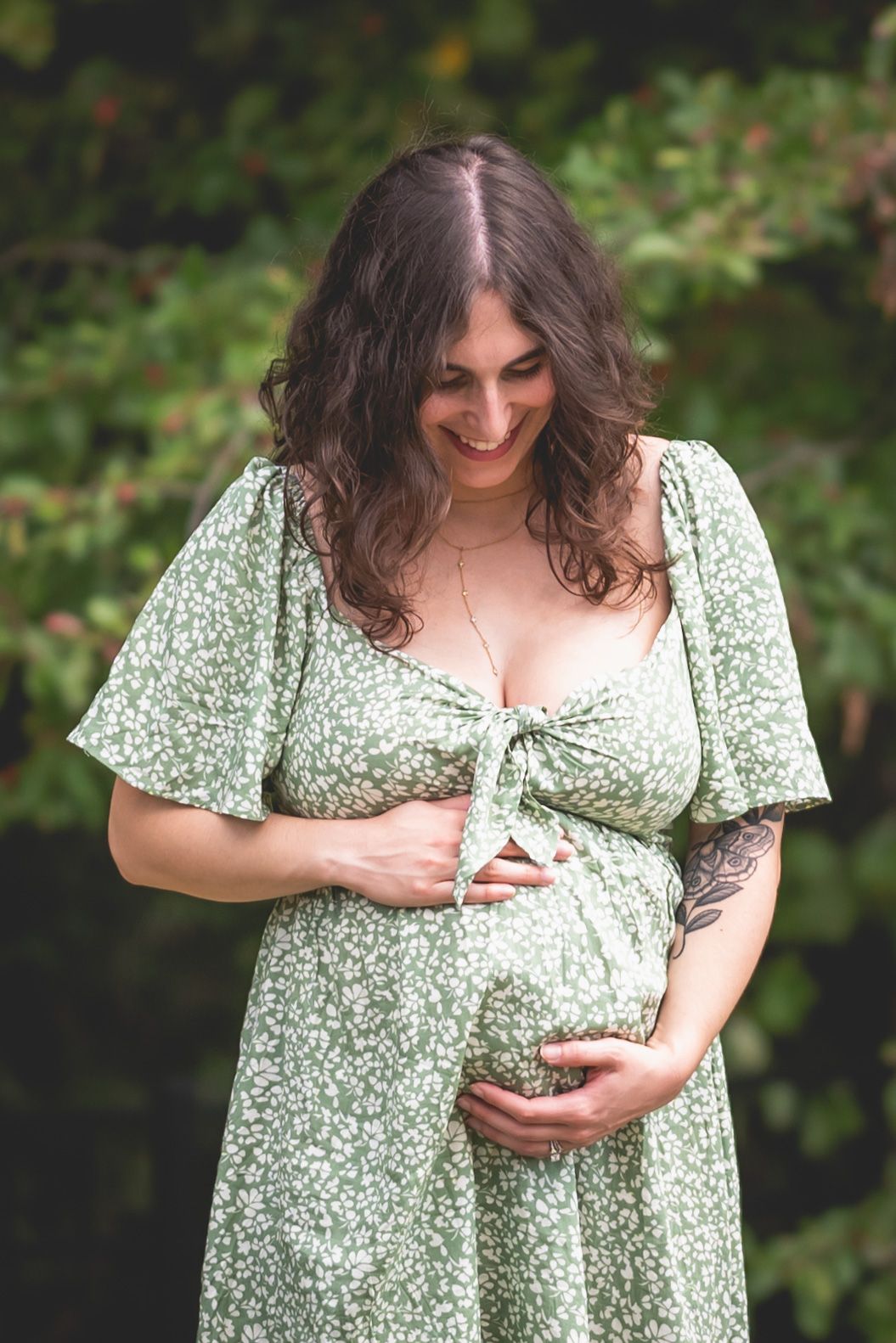 Smiling pregnant woman in green floral dress cradling baby bump outdoors among lush green foliage.