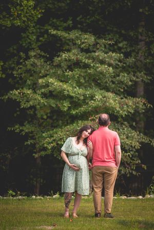 Pregnant couple sharing a tender moment outdoors, woman in green floral dress cradling baby bump, lush trees in background.