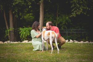 A couple kneels on grass petting a tall greyhound dog outdoors near trees and rocks.