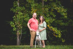 Couple with white dog posing outdoors on green grass with lush trees in background.