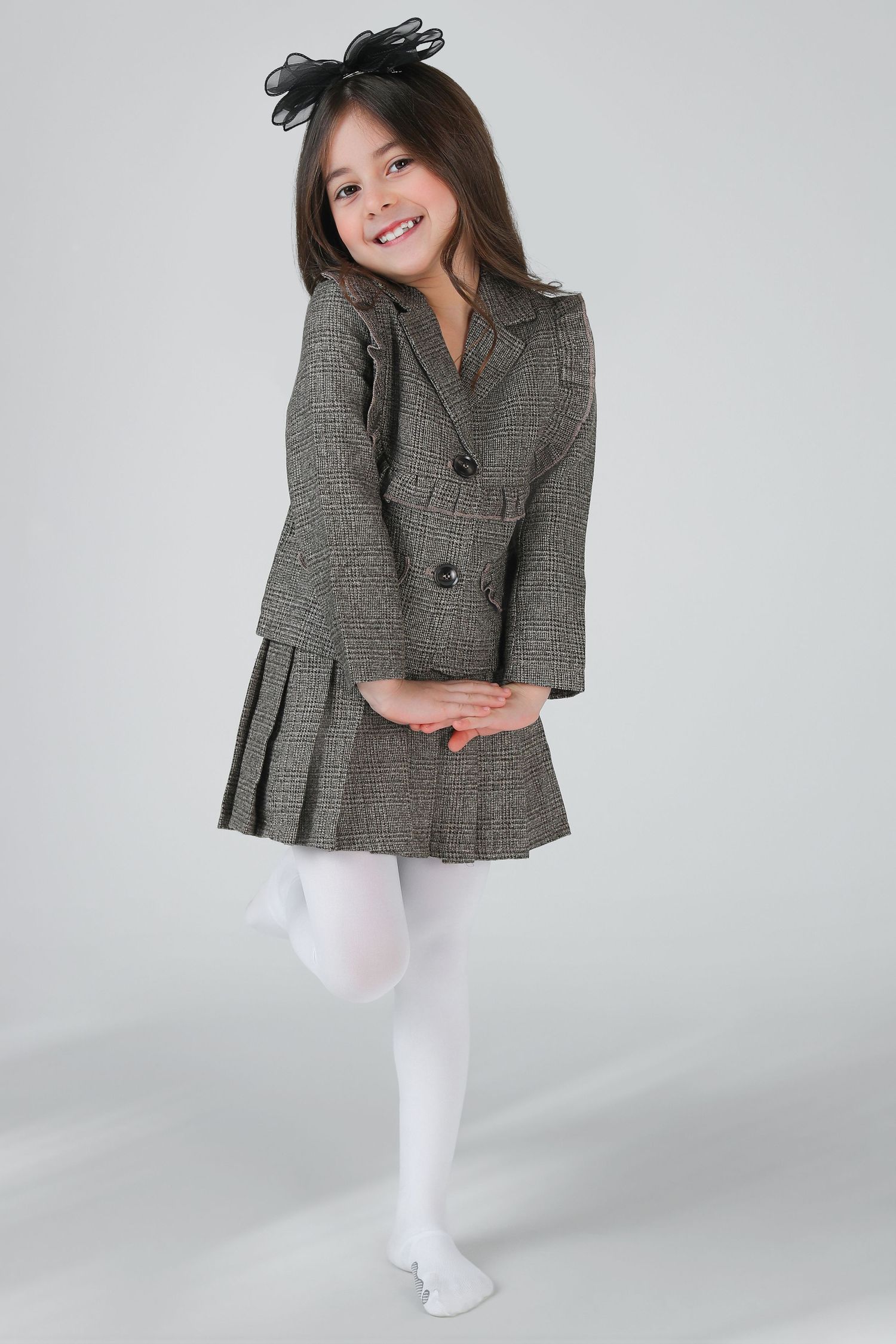 Young girl in plaid blazer and skirt with white tights and black bow headband, posing playfully.