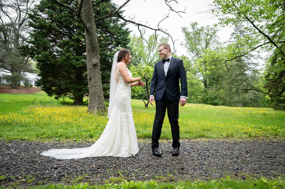Couple in wedding attire walking through lush green gardens with trees and yellow wildflowers in the background.