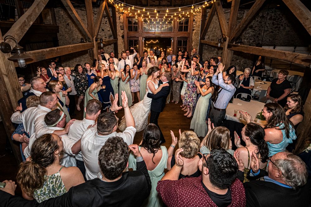 A lively crowd dances under string lights at a rustic barn wedding reception.