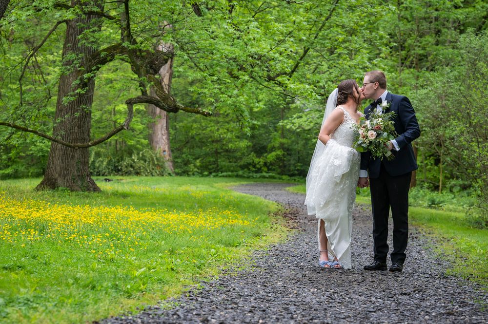 A couple walks down a winding gravel path surrounded by lush green trees and bright yellow wildflowers in a park.