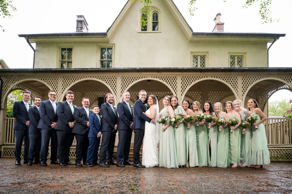 Wedding party members line up under a covered porch in coordinating formal attire for a panoramic group photo.