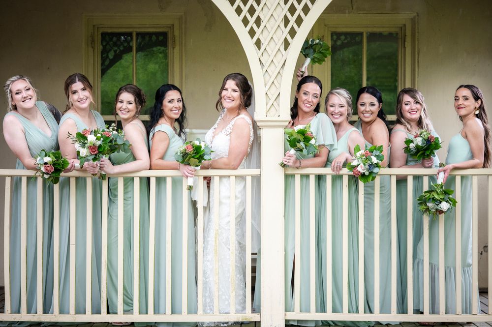 A row of bridesmaids in pink dresses stand on a white porch railing holding green bouquets.