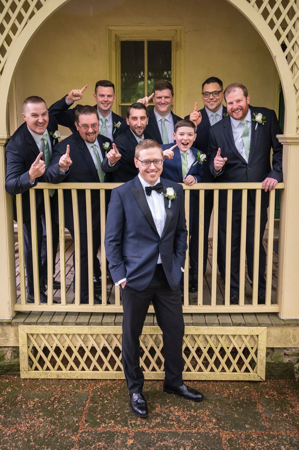 Groomsmen in navy suits pose playfully on a vintage porch with lattice details and autumn leaves on the ground.