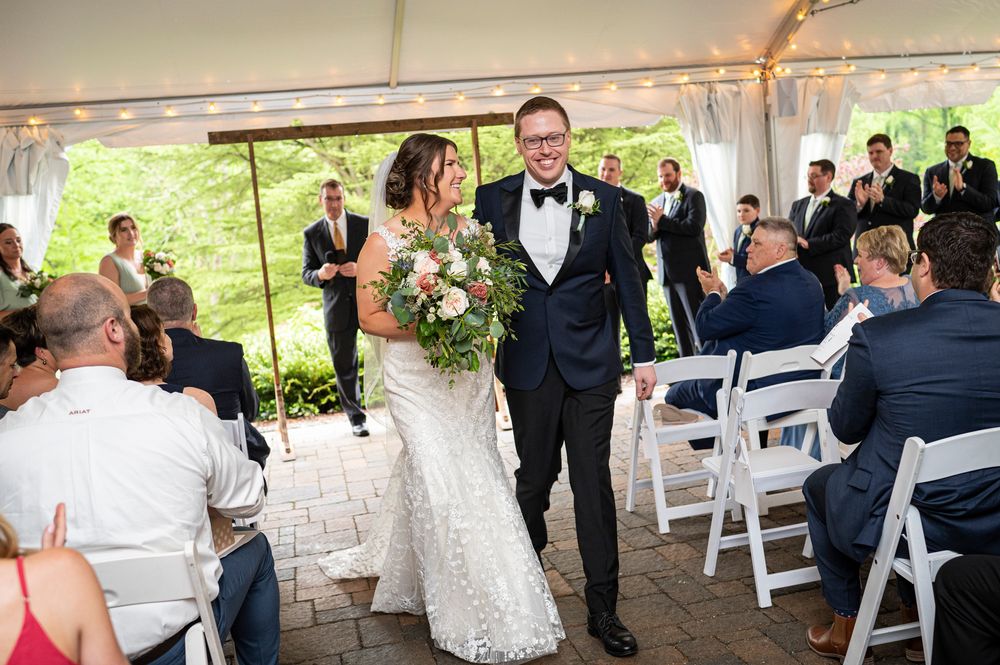 Wedding celebration under a white tent with guests seated at tables and white folding chairs.