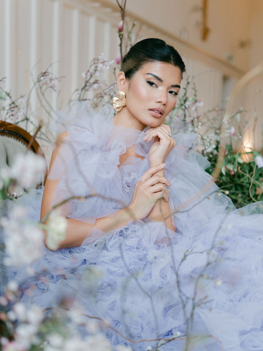 Model in ethereal lavender tulle gown poses elegantly among delicate floral arrangements in a romantic indoor setting.