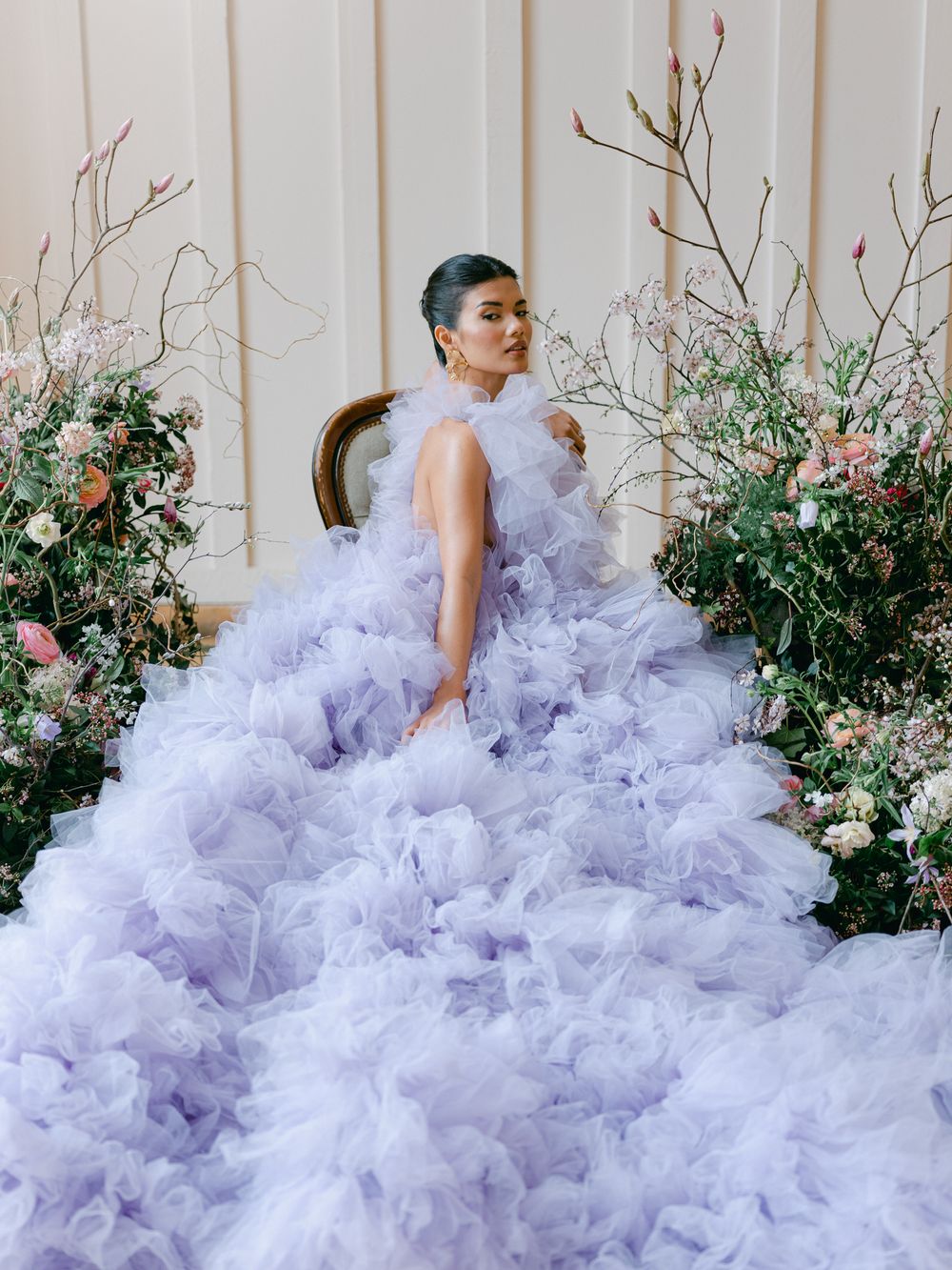 A dramatic fashion photo featuring a lavender tulle gown with voluminous ruffles against a floral garden backdrop.
