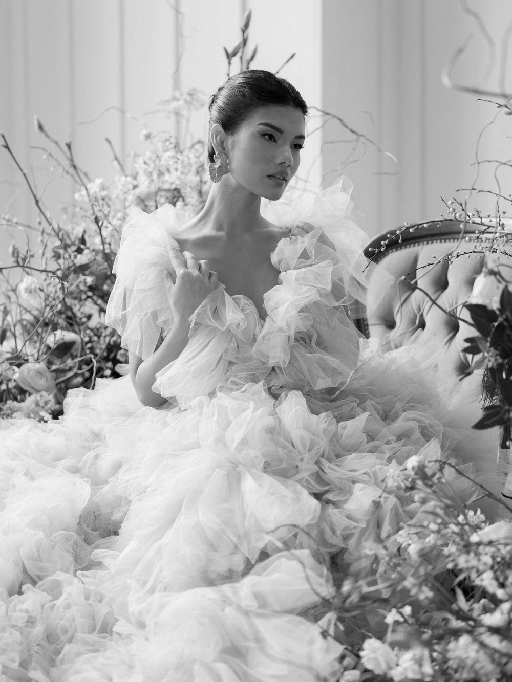 Elegant black and white fashion photograph featuring a dramatic white ruffled gown surrounded by delicate floral arrangements.