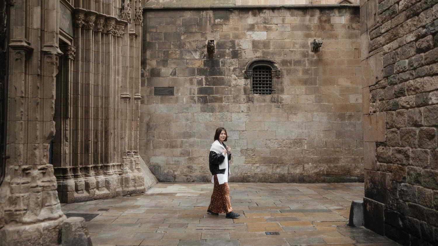 Woman in white coat and red pants walking beside ancient stone cathedral walls on a cloudy day.