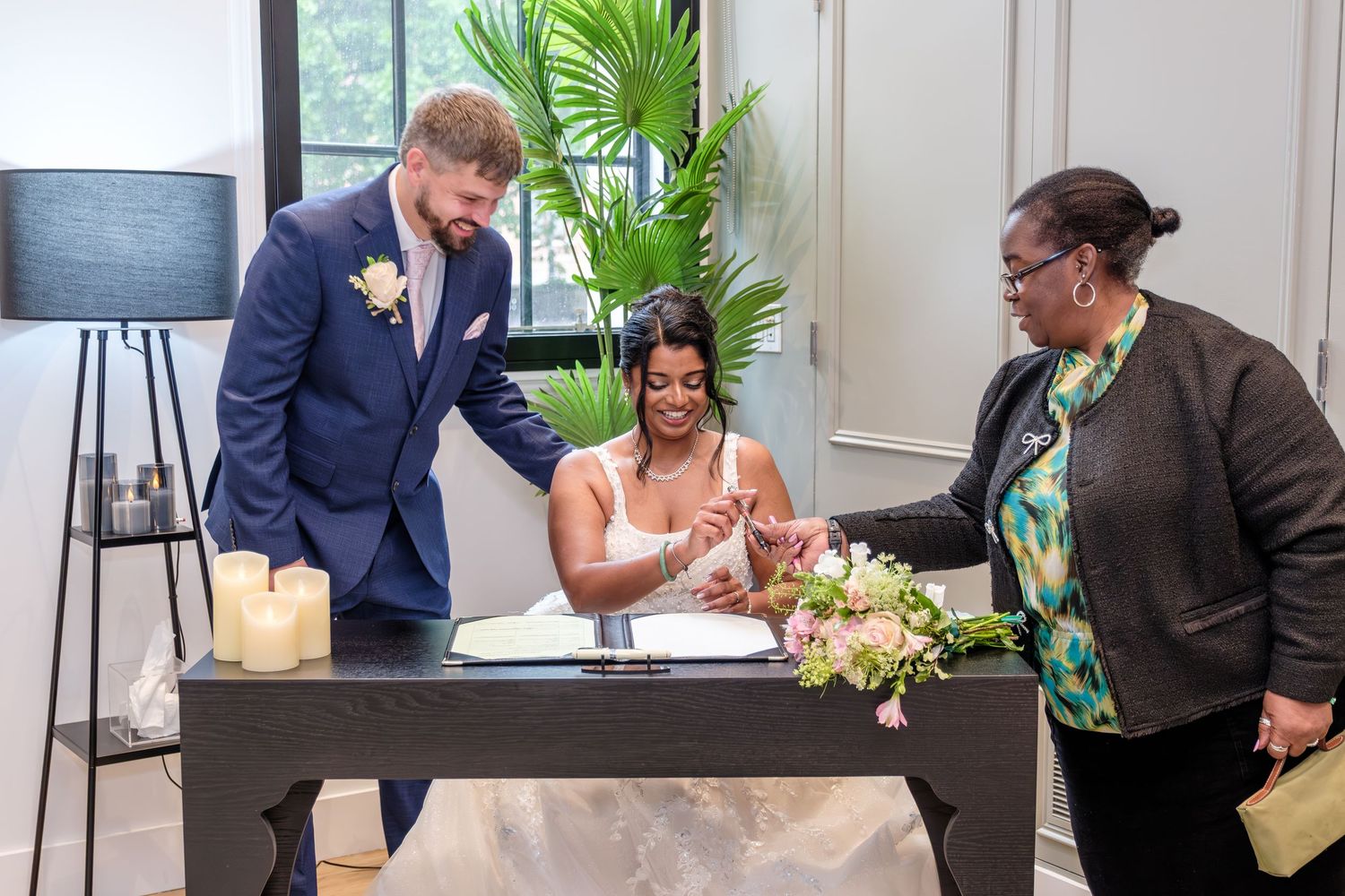 A bride and groom stand together surrounded by wedding guests in a formal indoor setting.