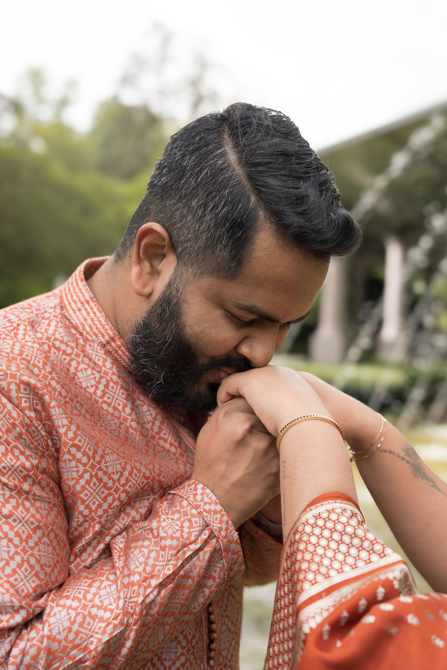 A bearded Indian man in an orange kurta gently kisses a woman's hand adorned with gold bangles outdoors.