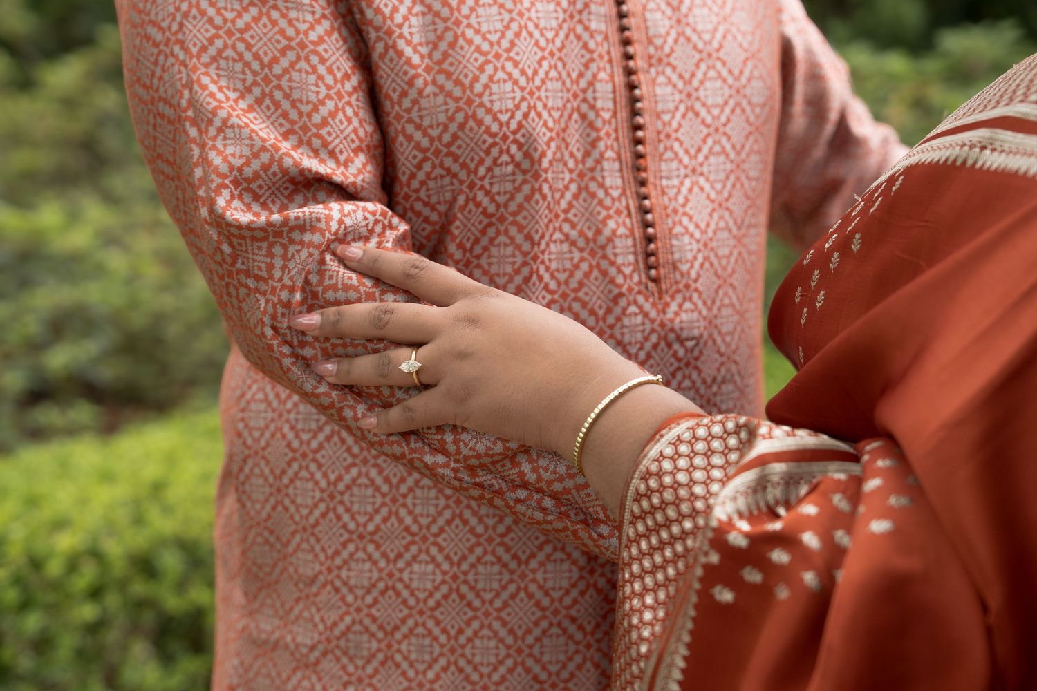 Close-up of a couple in traditional Indian attire, woman's hand with gold bracelet and ring resting on man's arm.