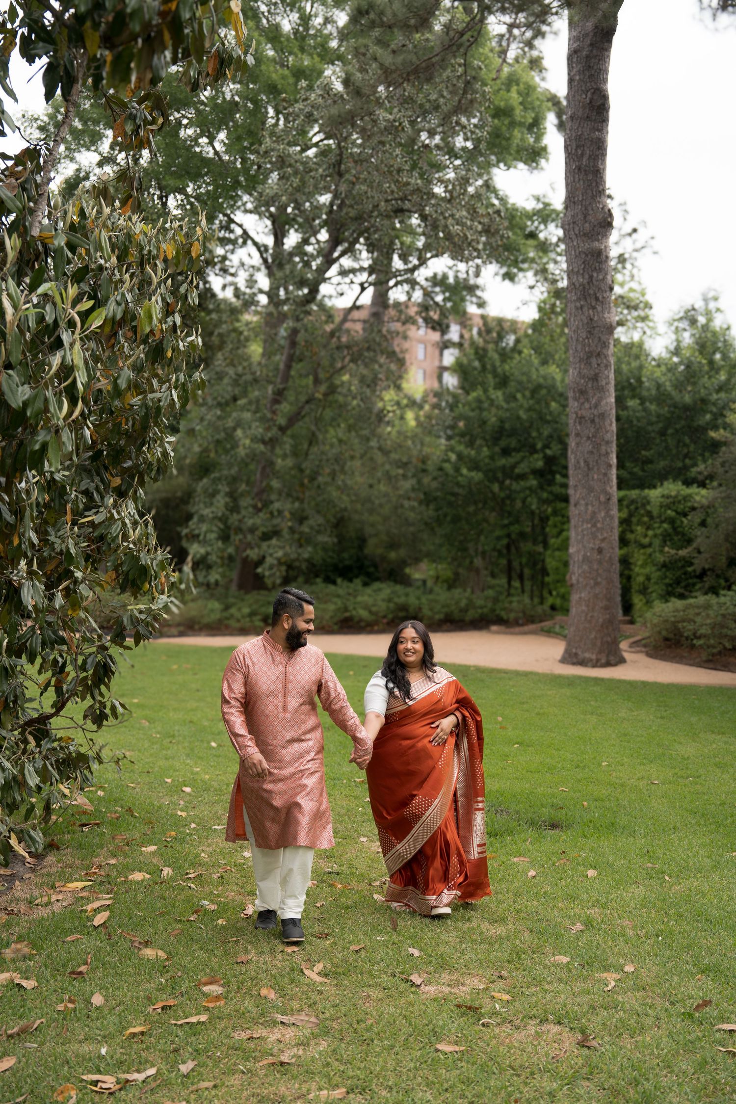 Indian couple in traditional attire walking hand in hand through a lush green park, smiling joyfully.