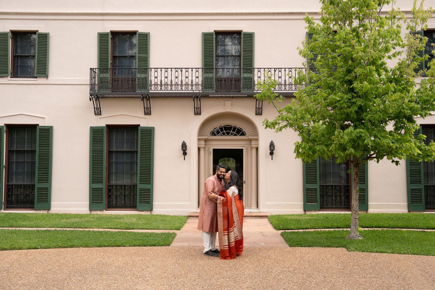 Indian couple in traditional wedding attire posing in front of an elegant white colonial mansion.