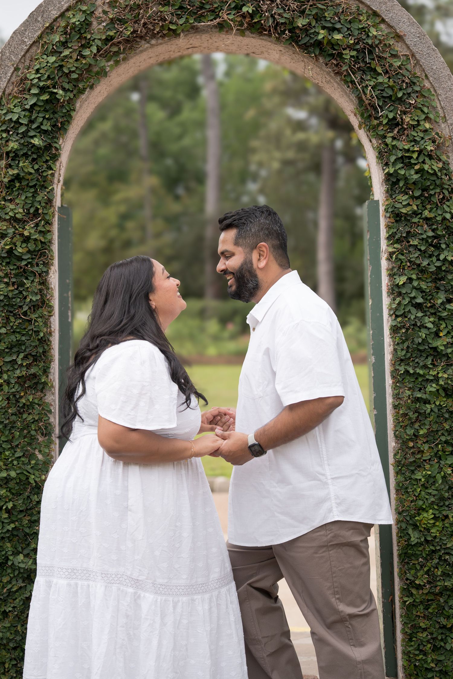 Couple holding hands and smiling at each other under an ivy-covered stone arch in a lush garden.