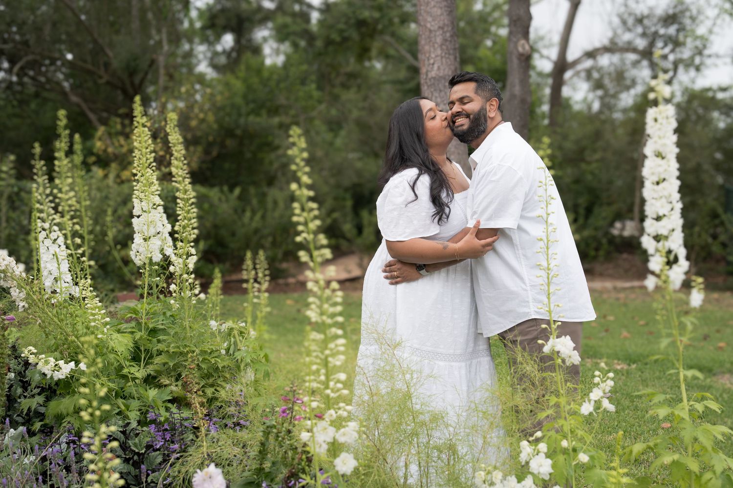 Couple in white outfits embracing among white flowering plants in lush garden.