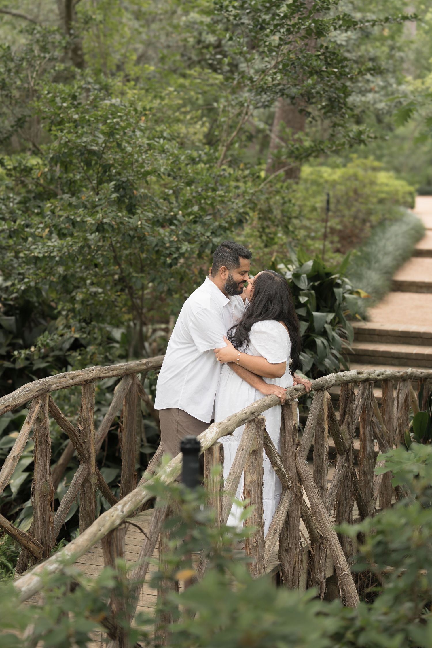 Romantic couple embracing and nearly kissing on a rustic wooden bridge surrounded by lush greenery.