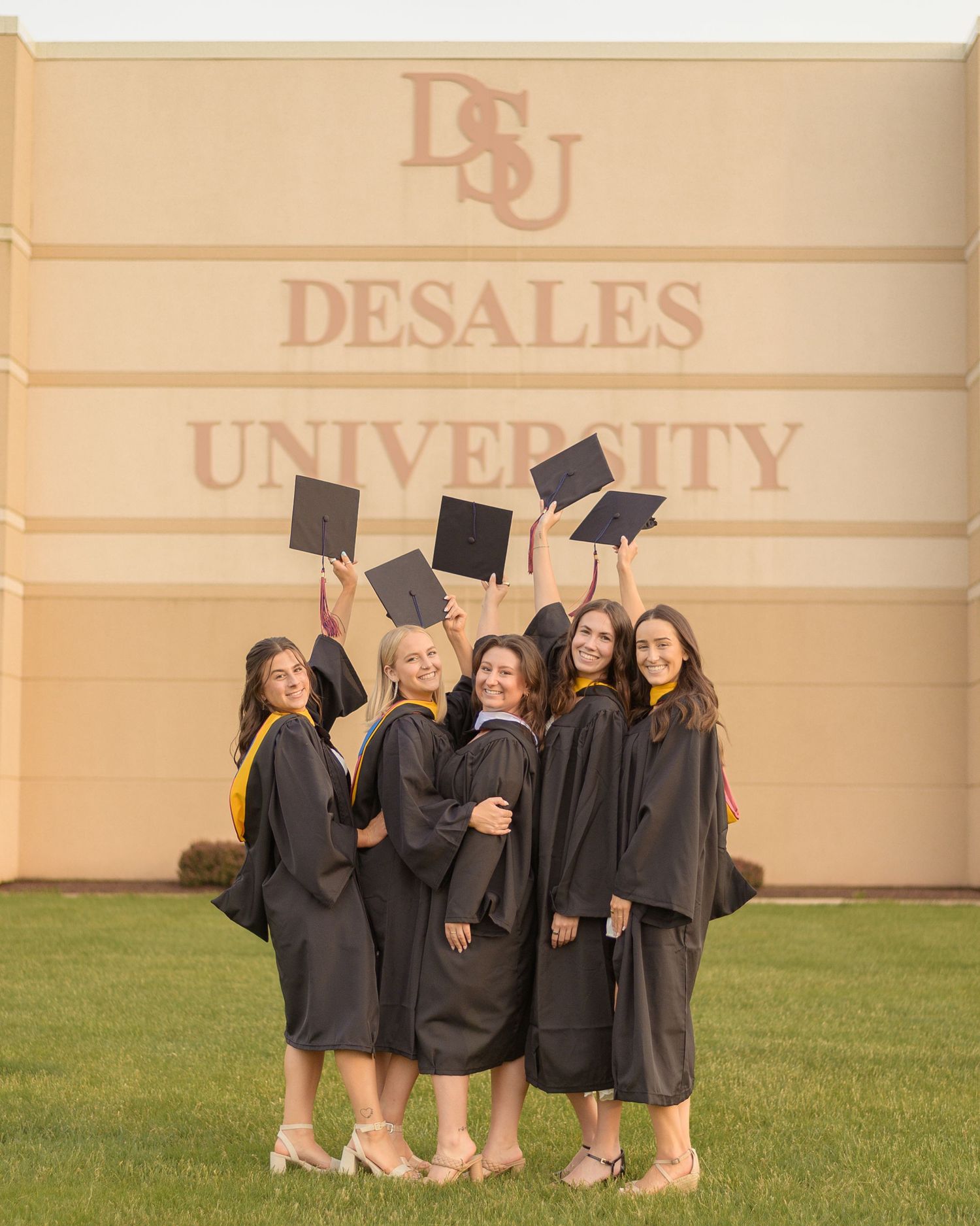 Graduates in black caps and gowns celebrate by tossing their mortarboards in front of DeSales University building.