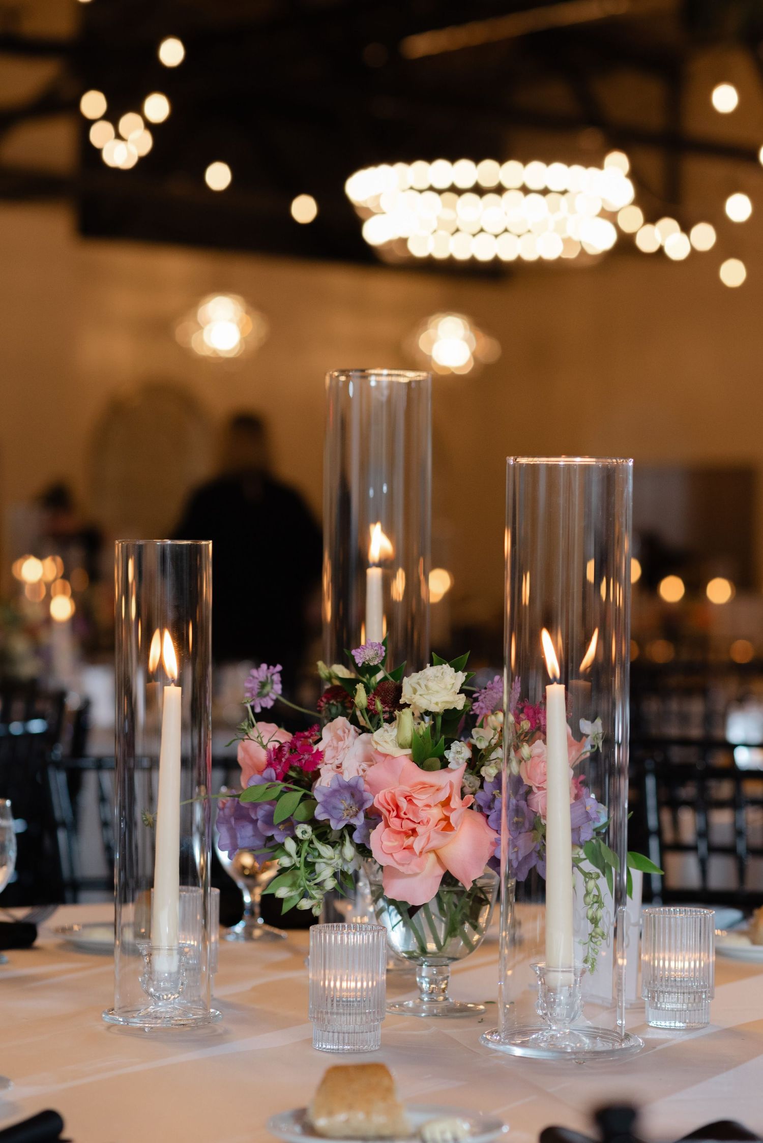 Elegant wedding centerpiece with pink and purple flowers surrounded by tall glass candle holders on white tablecloth.