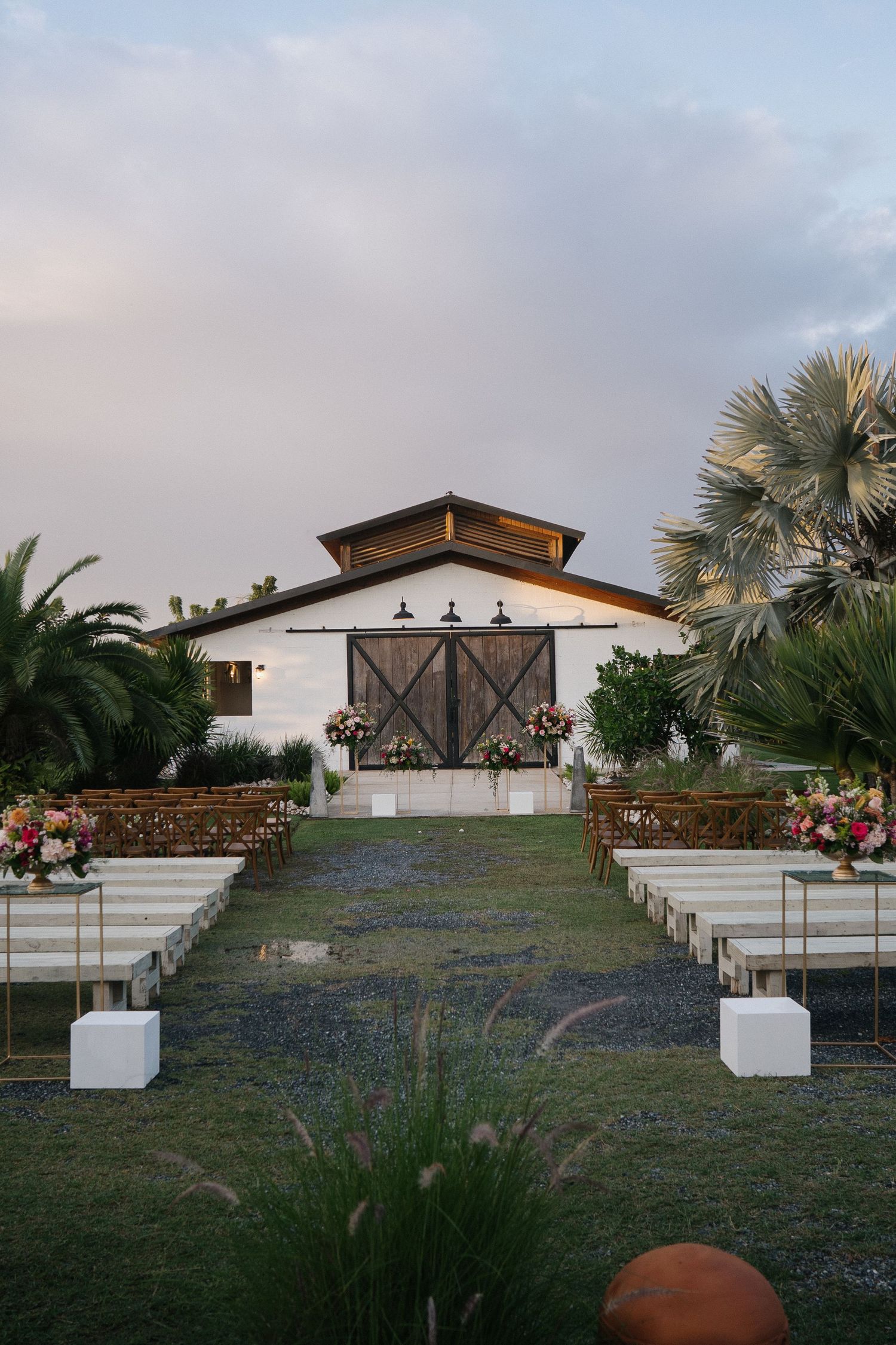 Rustic barn wedding venue with wooden doors and palm trees framing an outdoor ceremony aisle at dusk.