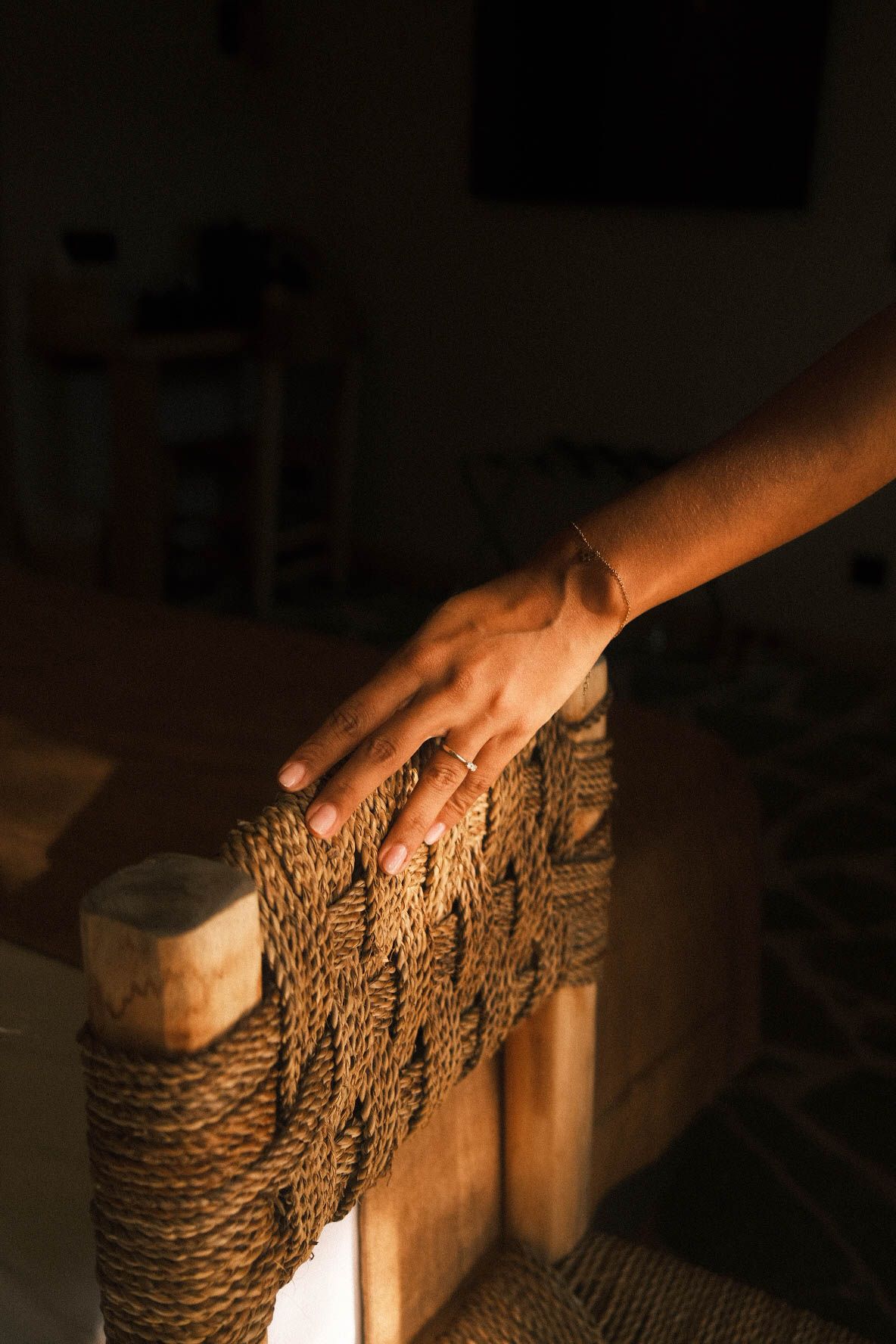 A hand with rings and bracelet rests on a woven rattan chair back in warm sunlight.