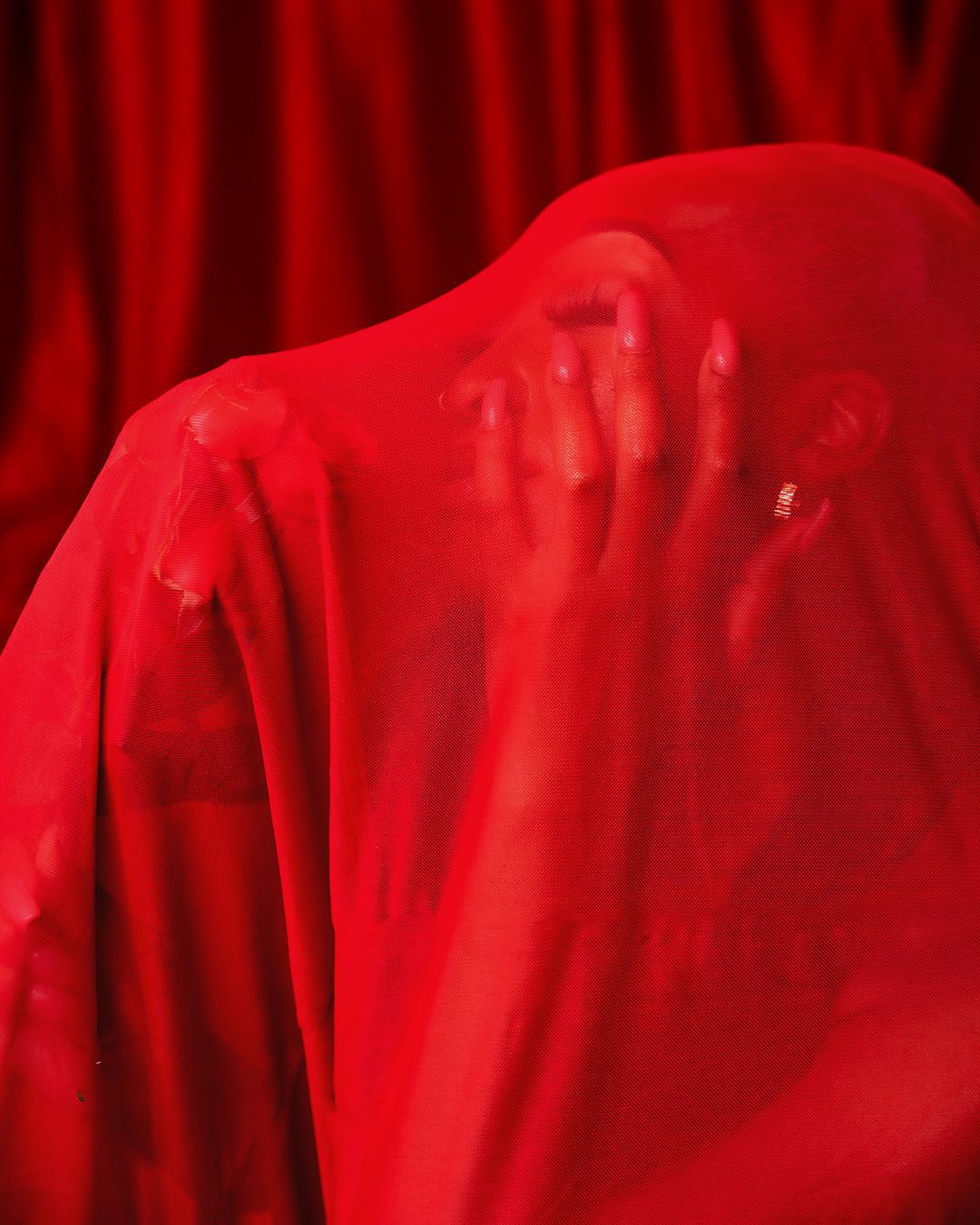Person draped in flowing red sheer fabric against red velvet curtains, hands visible through translucent material.