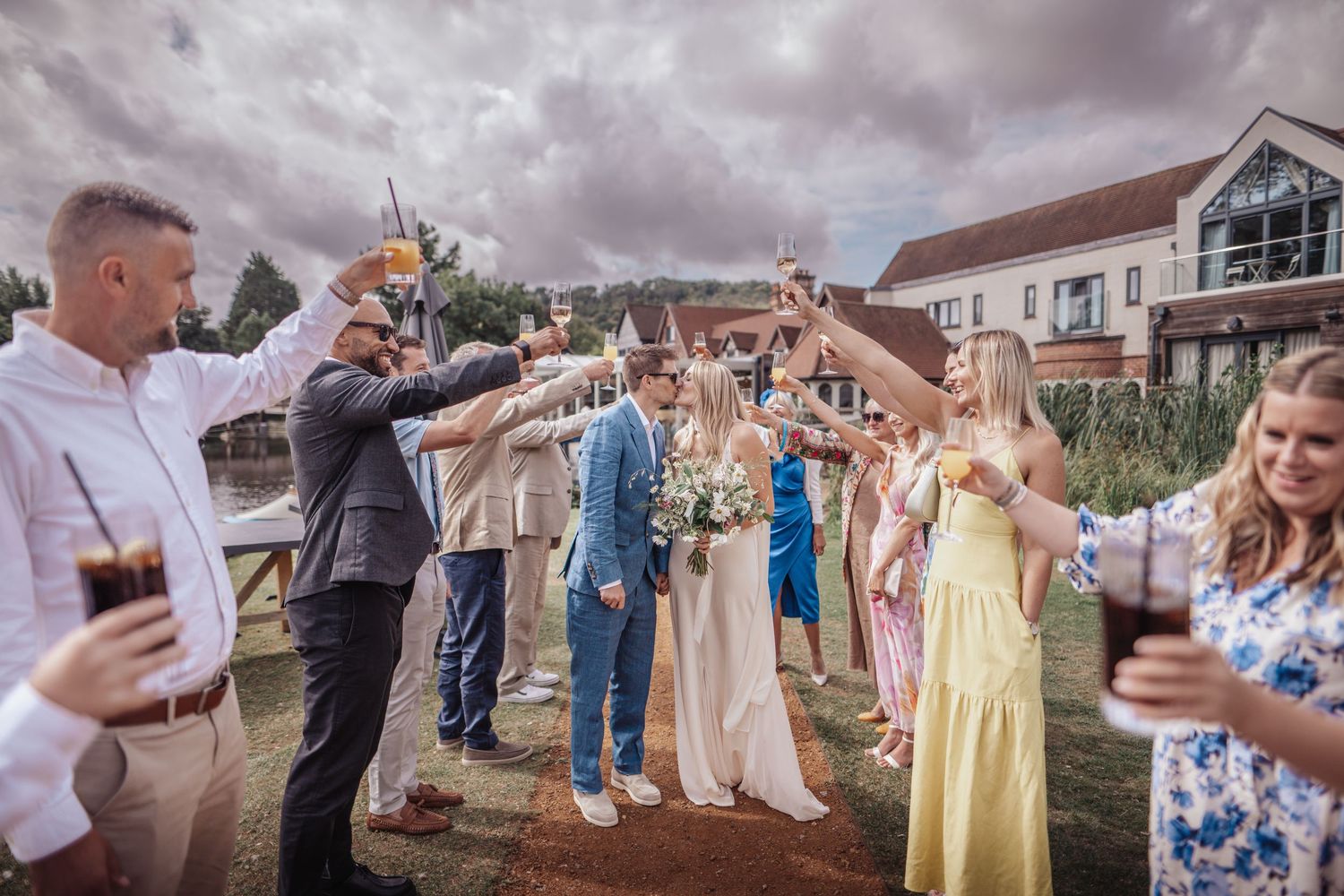Wedding guests raising glasses in a toast outdoors, with bride and groom at center, near a waterside venue.