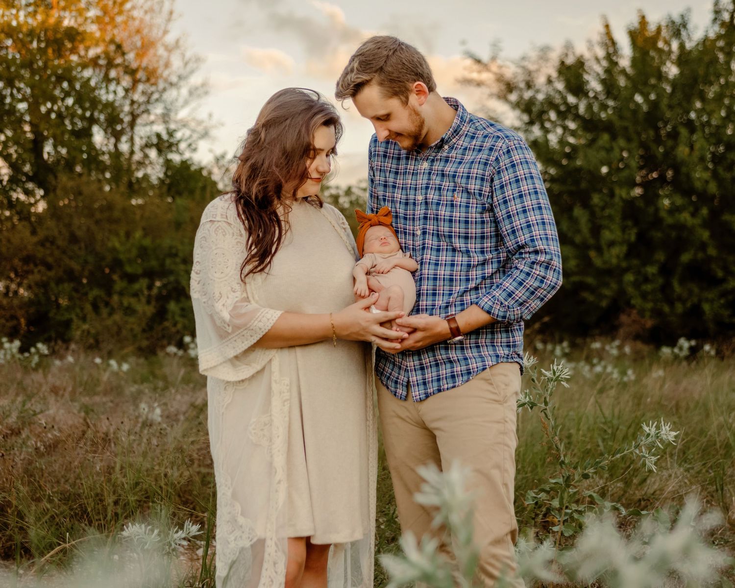 A couple holds their infant in a sunlit meadow during golden hour while wearing casual fall attire at their Fort Worth newborn photo session