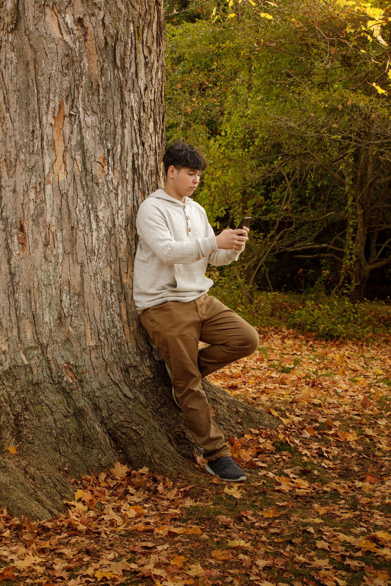 Person in white hoodie using phone while leaning against large tree trunk in autumn setting.