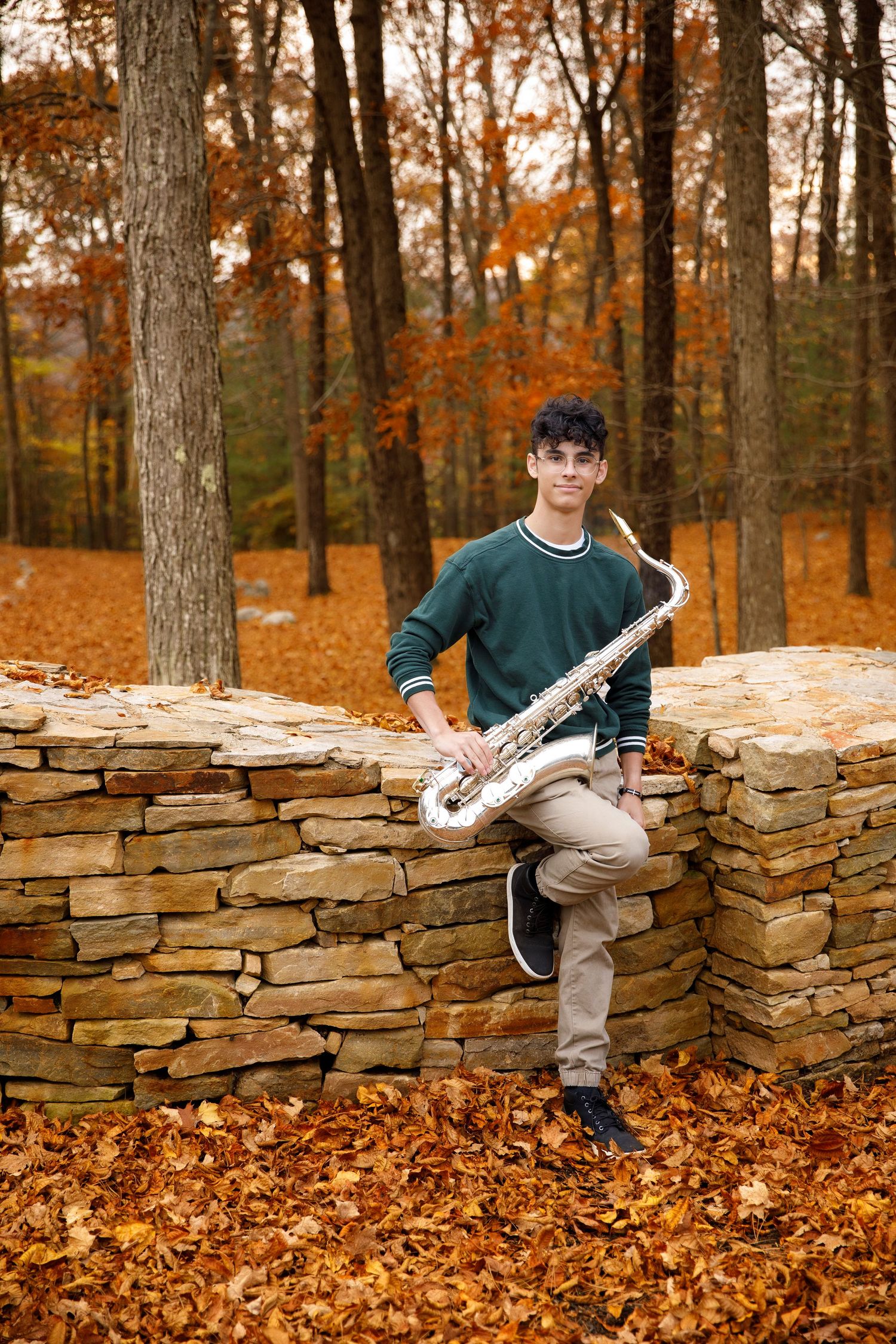 Musician with saxophone sits on stone wall surrounded by autumn foliage in forest setting.