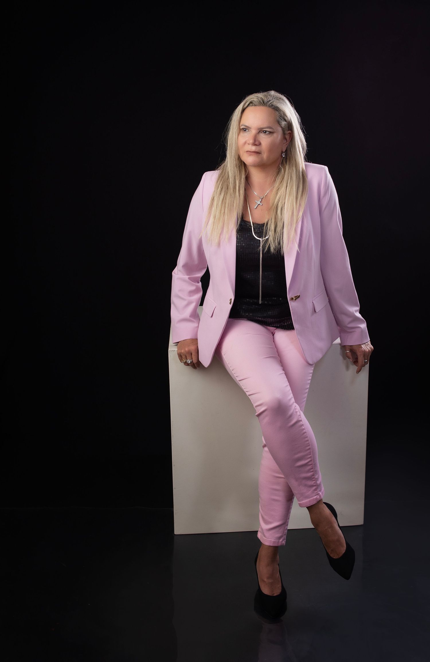 Professional wearing a pink business suit sits confidently on a white cube against dark background.