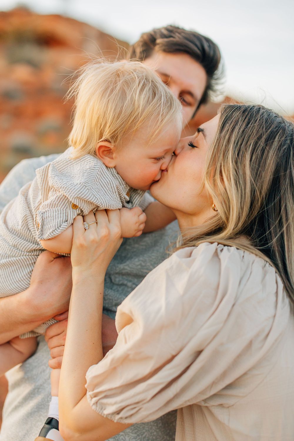 A woman tenderly kisses a baby while the man behind them embraces them both in a serene outdoor scene.