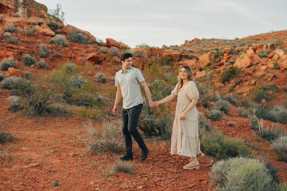 A couple stands together in a scenic desert landscape with red rock formations and vegetation.